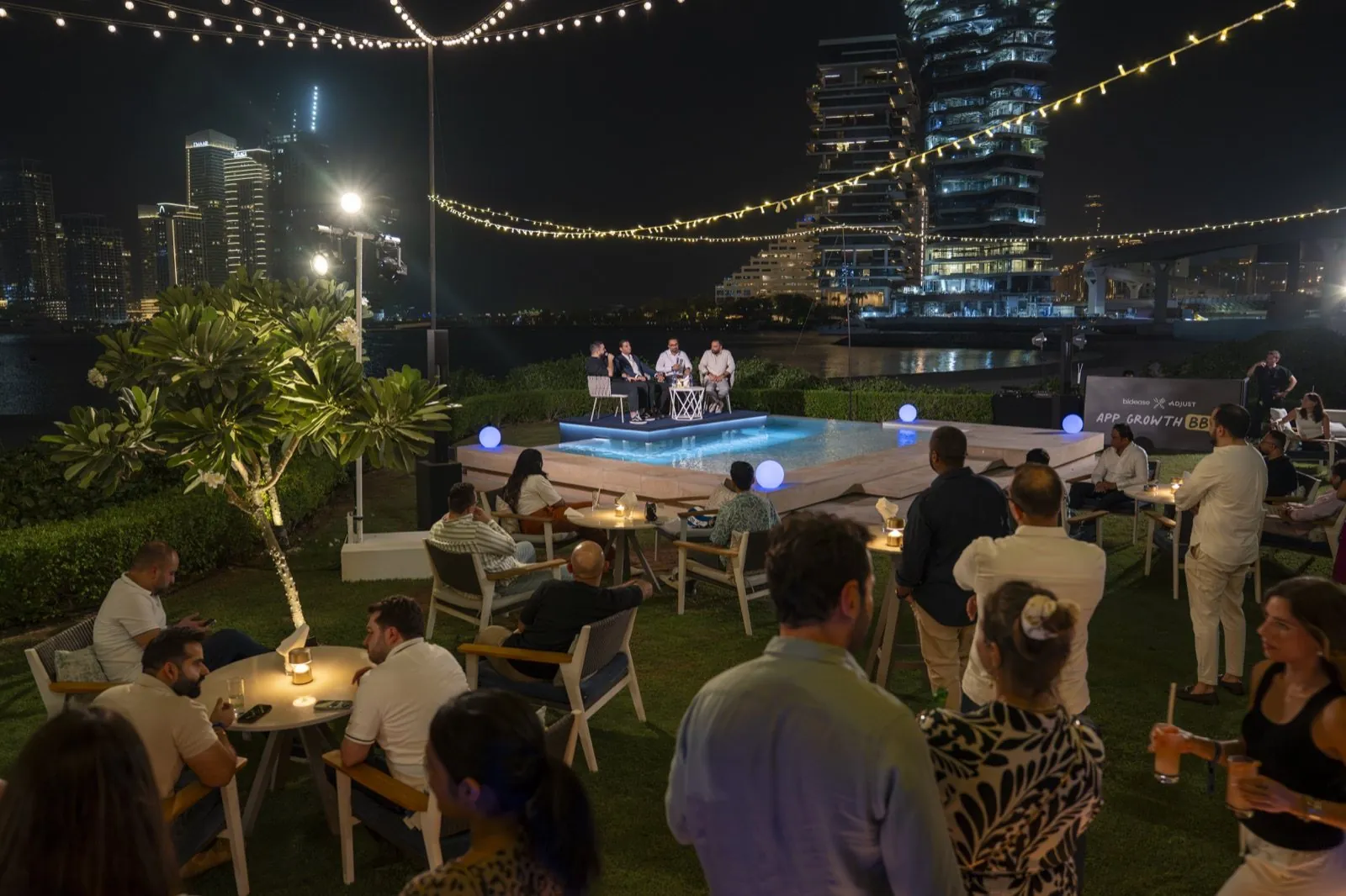 Evening garden event with a panel on a pool stage and the "bidease × Adjust App Growth BBQ" banner